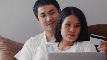 Young Asian Pregnant couple online shopping at home. Mom and Dad feeling happy using laptop technology and credit card buying baby product while lying on sofa in living room at home concept. photo