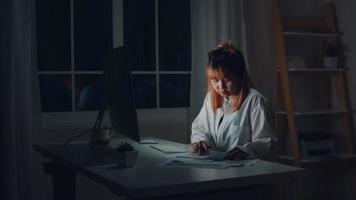 Young asian woman working late using desktop on desk in living room at home. Asia business woman writing notebook document finance and calculator in night at home office. Overworked female concept. photo
