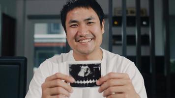 Portrait Asian Chinese man show and looking ultrasound photo baby in his wife at home. Young male relax toothy smile looking to camera sitting on table in living room at house in the morning concept.