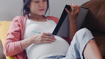 Young Asian Pregnant woman drawing baby in belly in notebook. Mom feeling happy smiling positive and peaceful while take care child lying on sofa in living room at home concept. photo