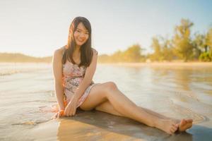 joven mujer asiática que se siente feliz en la playa, hermosa mujer feliz relajarse sonriendo diversión en la playa cerca del mar cuando se pone el sol por la noche. las mujeres de estilo de vida viajan en concepto de playa. foto