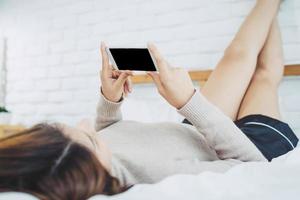 Happy Asian women are using smart phone with black blank empty screen on the bed in morning. Asian woman in bed checking social apps with smartphone. Smiling woman surfing net with cellphone at home. photo