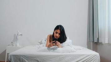 Young Asian woman using smartphone checking social media feeling happy smiling while lying on bed after wake up in the morning, Beautiful attractive hispanic lady smiling relax in bedroom at home. photo