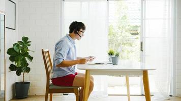 Asia businessman dressed in shirt and shorts use laptop talk to colleagues in video call while work from home at living room. Self-isolation, social distancing, quarantine for corona virus prevention. photo