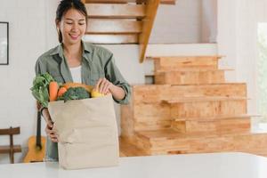 mujeres asiáticas que sostienen bolsas de papel para comprar comestibles en casa, una joven asiática feliz compra verduras y frutas, productos saludables y orgánicos del supermercado, póngalos en la cocina en el concepto de la mañana. foto