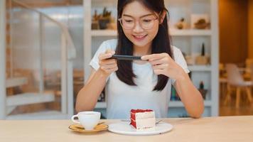 bloguera asiática amigable mujer influyente come pastel en el café de la noche. hermosa jovencita feliz relajarse diversión usando tecnología teléfono móvil tomando una foto de su carga de comida en las redes sociales en el campus universitario.