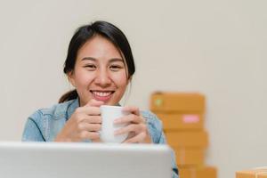 joven empresaria asiática propietaria de una pyme en línea que verifica el producto en stock, guarda en la computadora y toma café trabajando en casa. propietario de una pequeña empresa en el concepto de oficina en casa. foto