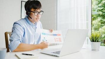 Young Asia businessman using laptop talk to colleagues about plan in video call while smart working from home at living room. Self-isolation, social distancing, quarantine for corona virus prevention. photo