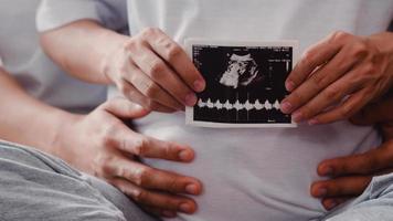 joven pareja asiática embarazada muestra y mira una foto de ultrasonido bebé en el vientre. mamá y papá se sienten felices sonriendo en paz mientras cuidan al niño acostado en el sofá en la sala de estar en el concepto de casa.