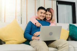 Attractive Asian sweet couple using computer or laptop while lying on the sofa when relax in their living room at home. Husband and his wife using relax and romantic time at home concept. photo