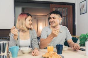 Happy sweet Asian couple having breakfast, cereal in milk, bread and drinking orange juice after wake up in the morning. Husband and his wife eating food together. photo