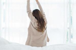 Back view of happy beautiful young Asian woman waking up in morning, sitting on bed, stretching in cozy bedroom, looking through window. Funny woman after wake up. She is stretching and smiling. photo
