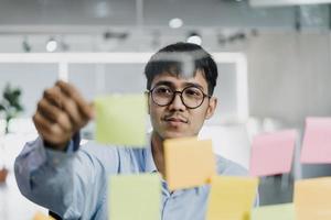 Asian creative man stick a sticky note on glass board. Young professional business male write information, strategy, reminder on paper, business situation, startup in Loft office concept. photo