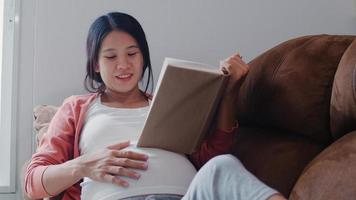 Young Asian Pregnant woman read a book for baby in belly. Mom feeling happy smiling positive and peaceful while take care child lying on sofa in living room at home concept. photo