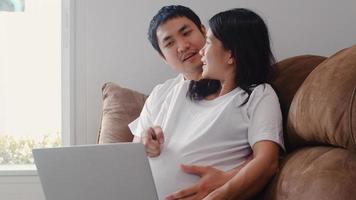 Young Asian Pregnant couple using laptop search pregnancy information. Mom and Dad feeling happy smiling positive and peaceful while take care their child lying on sofa in living room at home concept. photo