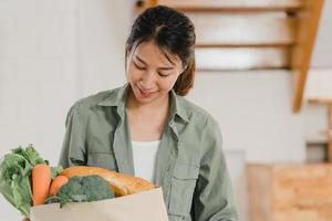 mujeres asiáticas que sostienen bolsas de papel para comprar comestibles en casa, una joven asiática feliz compra verduras y frutas, productos saludables y orgánicos del supermercado, póngalos en la cocina en el concepto de la mañana. foto