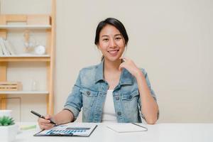 mujer de negocios de asia que se siente feliz sonriendo y mirando a la cámara mientras se relaja en la oficina en casa. joven asiática trabajando escribiendo finanzas de documentos de cuaderno en el escritorio en la sala de estar en casa.