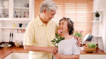 una pareja de ancianos asiáticos se siente feliz sonriendo y sosteniendo una flor y mirando a la cámara mientras se relaja en la cocina de casa. la familia mayor de estilo de vida disfruta del concepto de tiempo en casa. retrato mirando a la cámara.