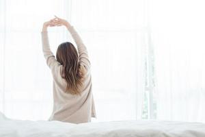 Back view of happy beautiful young Asian woman waking up in morning, sitting on bed, stretching in cozy bedroom, looking through window. Funny woman after wake up. She is stretching and smiling. photo