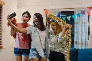 Group of Asian women party at home, female using mobile phone making vlog and selfie to social media while funny moment dancing together in living room in night. Young friend celebrate holiday concept photo