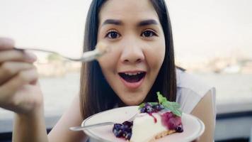 Blogger Asian friendly woman influencer eating cake at cafe. Young lady happy relax fun using technology mobile phone record vlog video upload in social media holiday travel near river in city. photo