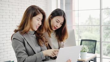 Two Asian young creative businesswomen holding a cup of coffee, working on laptop and disscuss about work while working in office. Casual business, freelance work at cafe, social meeting concept. photo