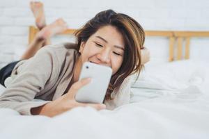 Happy Asian women are using smart phone on the bed in morning. Asian woman in bed checking social apps with smartphone. Smiling woman surfing net with cellphone at home. Mobile addict concept. photo