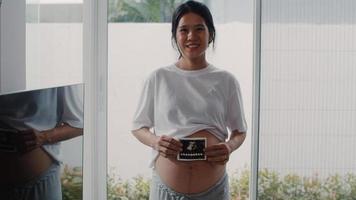Young Asian Pregnant women show and looking ultrasound photo baby in belly. Mom feeling happy smiling peaceful while take care child lying near window in living room at home concept.