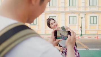 Traveler Asian couple travel in Bangkok, Thailand, sweet couple using mobile phone taking photo each other near street while spending holiday trip in sunset. Lifestyle couple travel in city concept.