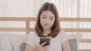 una joven asiática que usa un smartphone mientras está acostada en la cama después de despertarse por la mañana, una hermosa y atractiva chica japonesa que sonríe y se relaja en el dormitorio de su casa. disfrutando del concepto de estilo de vida de las mujeres en el hogar. foto