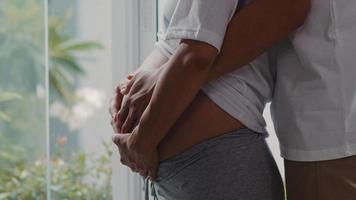 Young Asian Pregnant couple hug and holding belly talking with their child. Mom and Dad feeling happy smiling peaceful while take care baby, pregnancy near window in living room at home concept. photo