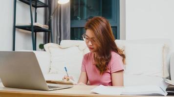 Young Asia teen girl student distance learning lesson with online teacher and study on computer laptop in living room from home at night. Social distancing, quarantine for corona virus prevention. photo