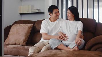 Young Asian Pregnant couple man touch his wife belly talking with his child. Mom and Dad feeling happy smiling peaceful while take care baby, pregnancy lying on sofa in living room at home concept. photo