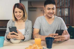 Atractiva pareja asiática joven distraída en la mesa con el periódico y el teléfono celular mientras desayuna. Emocionada joven pareja asiática sorprendida por una increíble buena noticia, familia feliz asombrada por Internet. foto