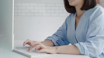 hermosa joven sonriente mujer asiática que trabaja en la computadora portátil mientras está en casa en el espacio de trabajo de oficina. empresaria trabajando desde casa a través de un ordenador portátil escribiendo en el teclado. disfrutando del tiempo en casa. foto