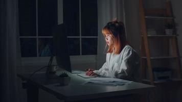 Young asian woman working late using desktop on desk in living room at home. Asia business woman writing notebook document finance and calculator in night at home office. Overworked female concept.