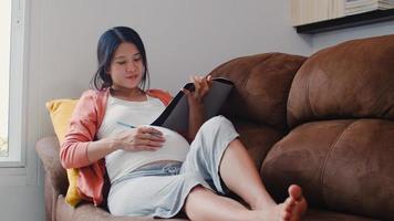 Young Asian Pregnant woman drawing baby in belly in notebook. Mom feeling happy smiling positive and peaceful while take care child lying on sofa in living room at home concept. photo
