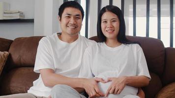 Young Asian Pregnant couple making heart sign holding belly. Mom and Dad feeling happy smiling peaceful while take care baby, pregnancy lying on sofa in living room at home concept. photo