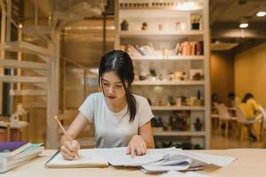 Asian student women reading books in library at university. Young undergraduate girl do homework, read textbook, study hard for knowledge on lecture desk at college campus overtime night. photo