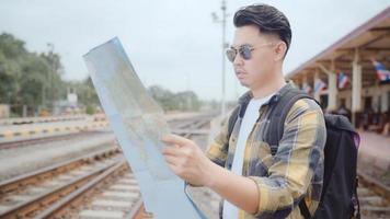 Traveler Asian man direction and looking on location map while spending holiday trip and waiting train at train station, Young male tourist backpacker enjoy journey. Lifestyle men travel concept.
