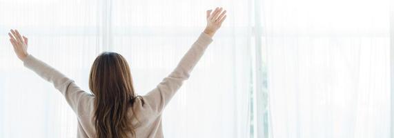 Back view of happy beautiful young Asian woman waking up in morning, sitting on bed, stretching in cozy bedroom. Funny woman after wake up. She is stretching and smiling. Panoramic banner. photo