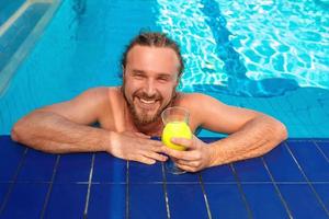 smiling cheerful caucasian european adult young man with dreadlocks and beard with cocktail near open swimming pool in summer outdoor. Vacation and sport concept. photo