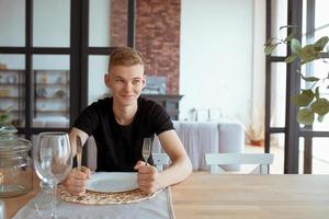 portrait of hungry handsome young man in black t-shirt sitting at the table with fork, knife and empty plate in loft interior indoor photo