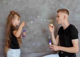 caucasian siblings - teenager boy brother and little girl sister with bubbles in modern loft interior on gray cement background photo