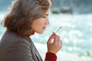 hermosa y elegante mujer joven estresante y triste con suéter y abrigo fumando cigarrillos cerca del río foto