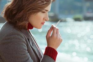 hermosa y elegante mujer joven estresante y triste con suéter y abrigo fumando cigarrillos cerca del río foto