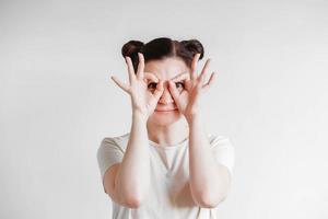 Portrait of a beautiful woman with her hands emitting glasses and a funny face wearing a T-shirt on a white background. Copy, empty space photo