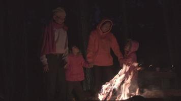 Family warms near campfire in forest and having a conversation. Night camping near bonfire in pine forest. Tourism and camping concepts.