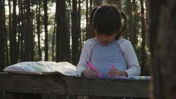 Little girl coloring pictures in a campsite in a pine forest. Creative leisure for little child. Children spend time with family on vacation.