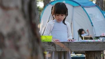 niña coloreando fotos en un camping en un bosque de pinos. Ocio creativo para niños pequeños. los niños pasan tiempo con la familia de vacaciones. video